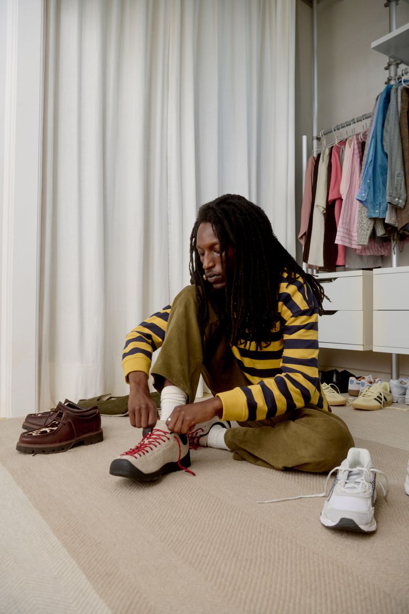 Model Eden Burundi tying cream sneakers with red laces, wearing a yellow striped sweater and olive pants in a wardrobe setting.