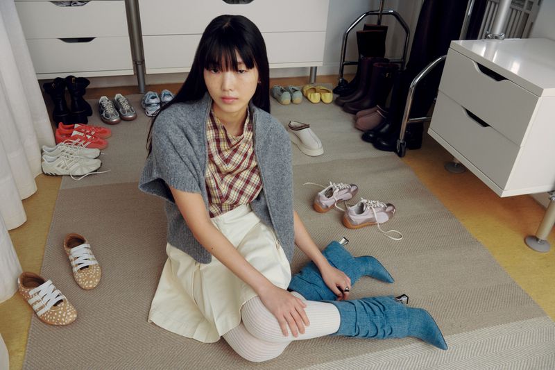 Model Hanna Jang sits on a carpet surrounded by pairs of sneakers and boots wearing a grey knit vest and denim slouch boots.