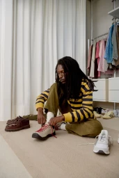 Model seated cross-legged on a carpeted floor tying the laces of a beige sneaker beside a shoe collection in a wardrobe room.