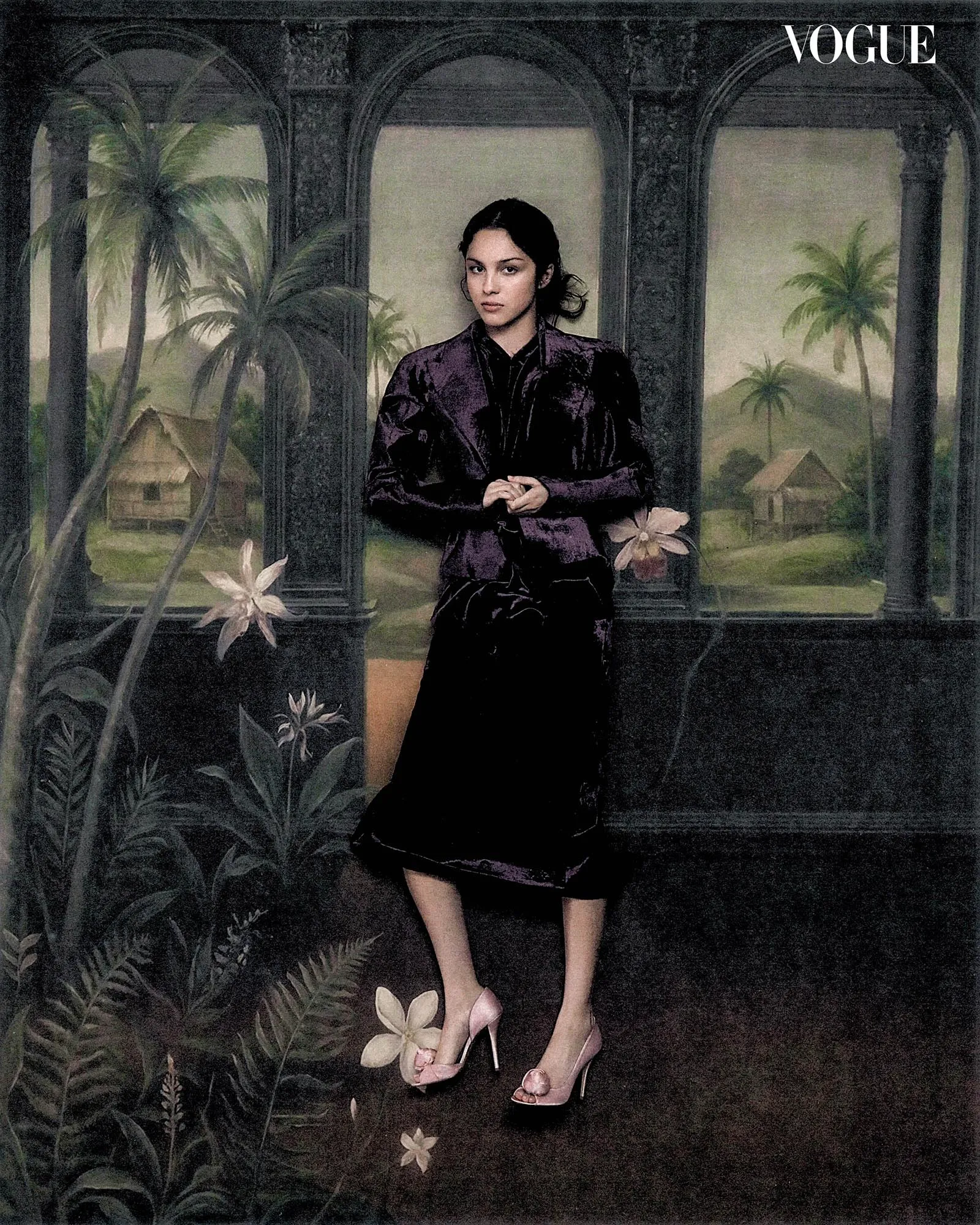 Olivia Rodrigo in a dark velvet jacket and matching skirt standing before a painted tropical mural for Vogue Philippines