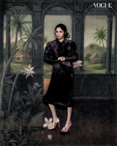 Olivia Rodrigo posing in a dark velvet skirt suit and pink heels in front of a painted tropical arch backdrop for Vogue Philippines.