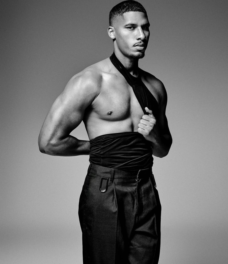 Black and white studio portrait of a muscular, shirtless adult male in dark dress pants, posing against a gray backdrop with a neutral expression.