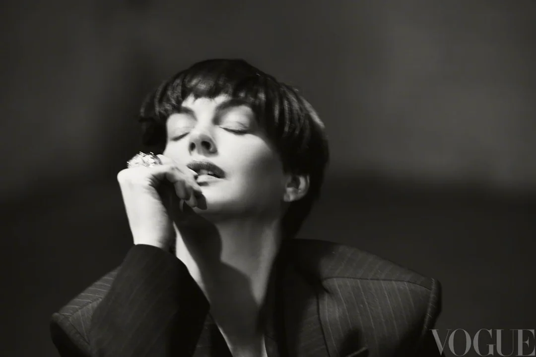 Black and white portrait of Anne Hathaway with short dark hair wearing a pinstripe blazer with her hand raised near her lips.