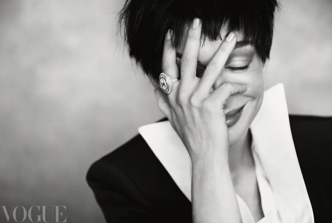 Anne Hathaway smiling while resting her hand on her face, wearing a statement ring and a black jacket with a prominent white collar.