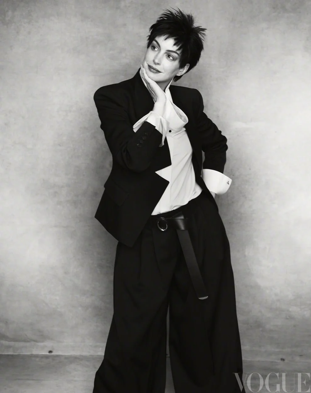 Anne Hathaway with short dark hair wearing an oversized black tailored suit and white shirt against a textured grey backdrop.