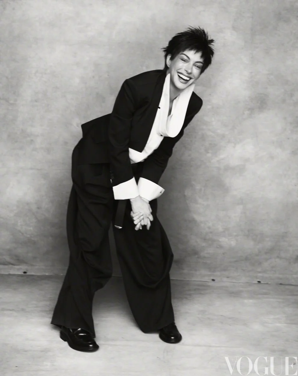 Anne Hathaway laughing in an oversized black suit and white collared shirt against a mottled studio backdrop.