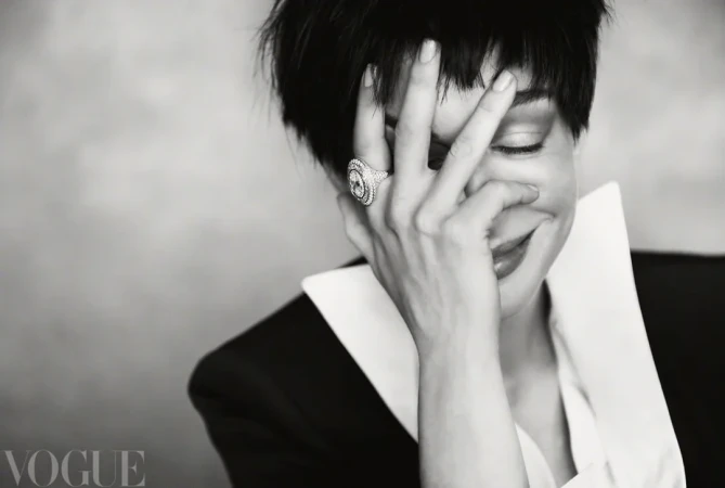 Anne Hathaway smiling while resting her hand on her face, wearing a statement ring and a black jacket with a prominent white collar.