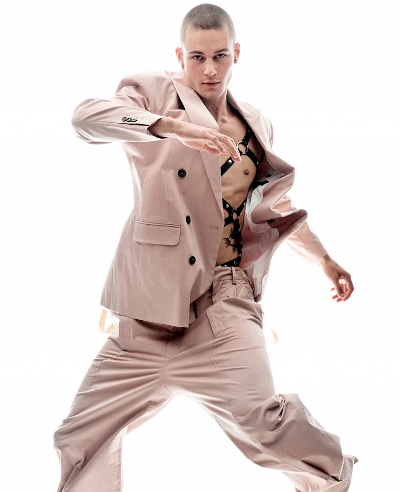 Man in a pink suit posing in a brightly lit studio. He is wearing a black leather harness and has a shaved head.