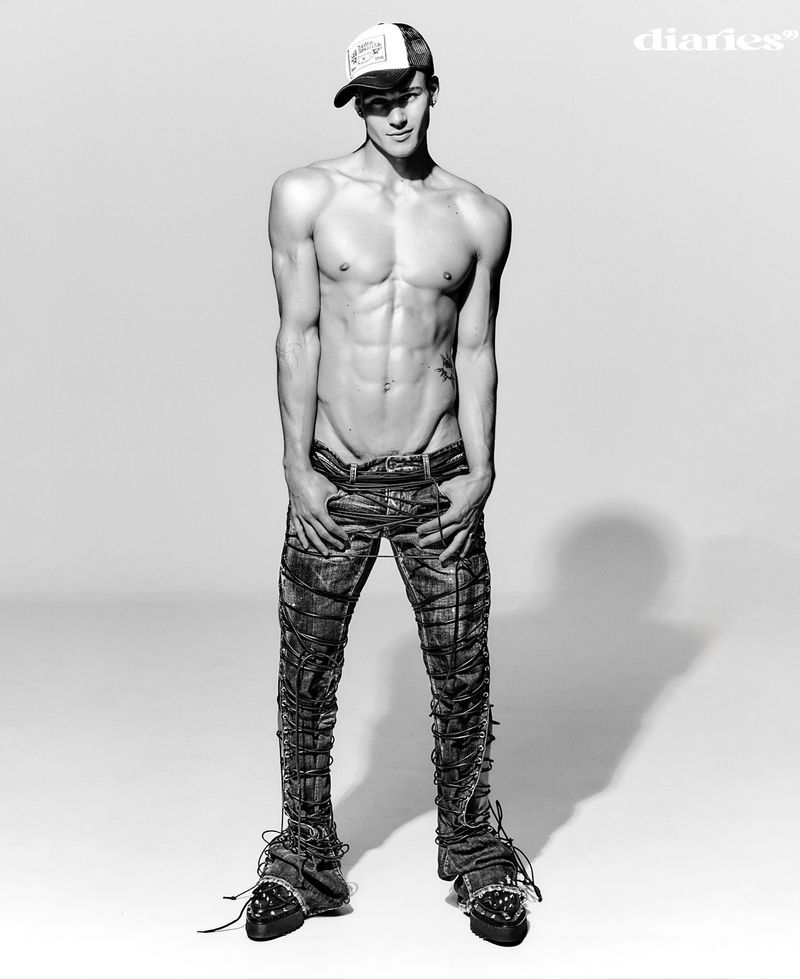 Black-and-white studio portrait of a muscular man in a baseball cap and textured jeans, striking a confident pose against a neutral backdrop.