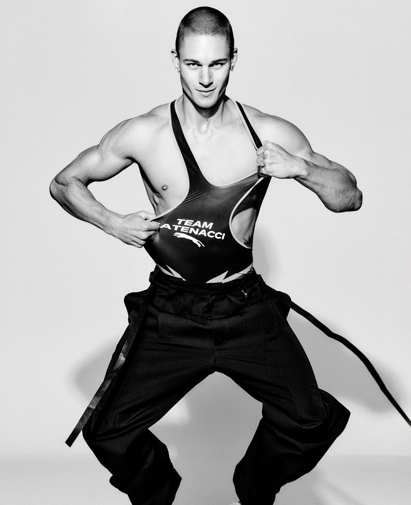 Black-and-white studio shot of muscular adult male lifting his athletic tank top, showcasing athletic physique, posing in a studio setting.