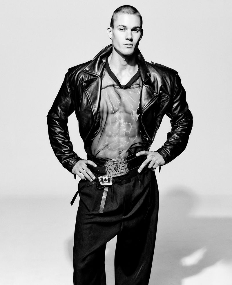 Studio shot of a young adult male with buzz cut hair in a sheer top, leather jacket, dark pants and statement belt against white.