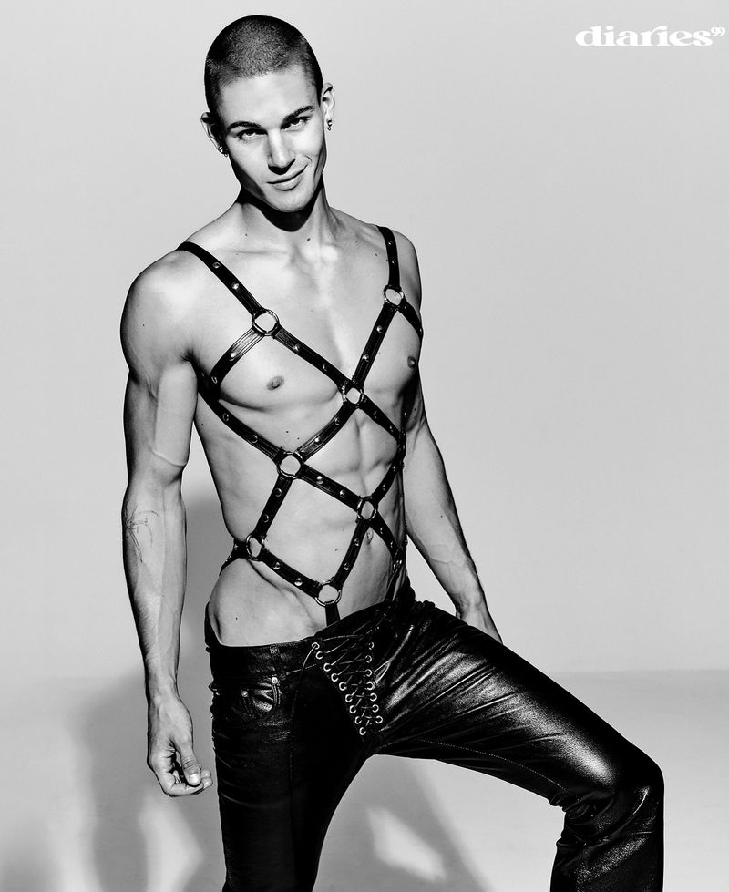 A muscular man in leather pants and a harness poses in a studio, showcasing a fashion-forward and edgy aesthetic with a confident expression.