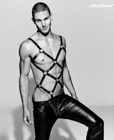 A muscular man in leather pants and a harness poses in a studio, showcasing a fashion-forward and edgy aesthetic with a confident expression.