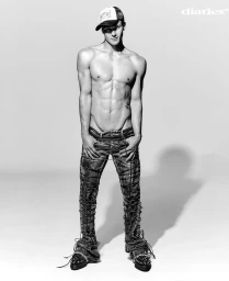 Shirtless male model wearing a cap and patterned denim trousers against a white studio background.