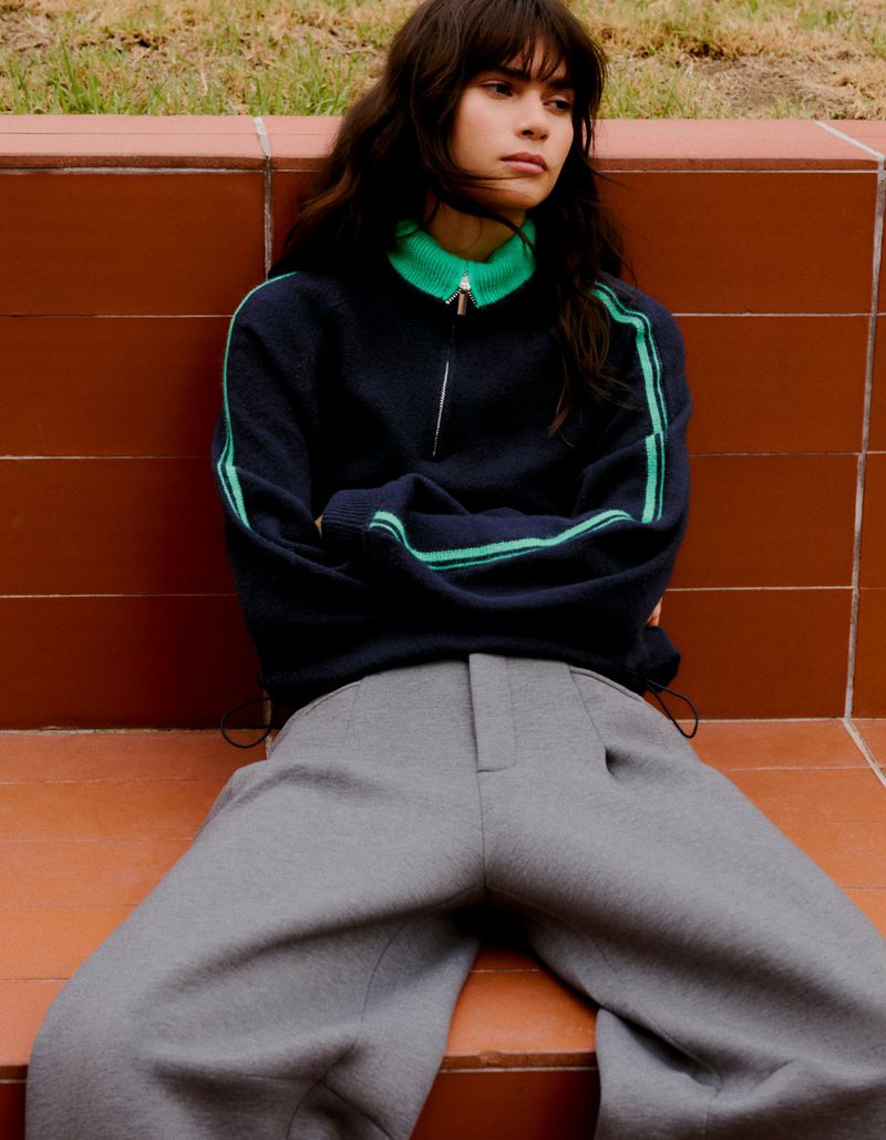 Model Clara Marques seated against terracotta tiles wearing a navy half-zip sweater with a green collar and grey wide-leg pants.