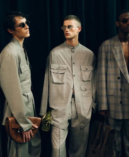 Three adult male fashion models pose indoors in a line, showcasing suits, shirts, and accessories against a dark, draped background.
