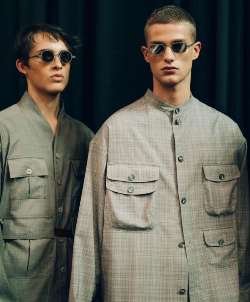 Two adult males in matching, neutral-toned button-down shirts with large pockets and retro sunglasses pose against a dark background.