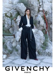 Model Kaia Gerber wearing a black blazer and wide-leg trousers standing before a rocky cliff face for Givenchy