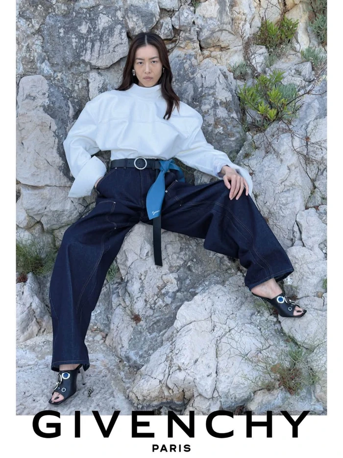 Model Liu Wen seated against a rocky stone wall wearing a white top and dark trousers for Givenchy