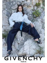 Model Liu Wen seated against a rocky stone wall wearing a white top and dark trousers for Givenchy
