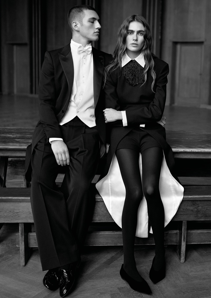 A man in a black tuxedo and white bow tie and a woman in a black tailored dress and tights seated together on a wooden bench.