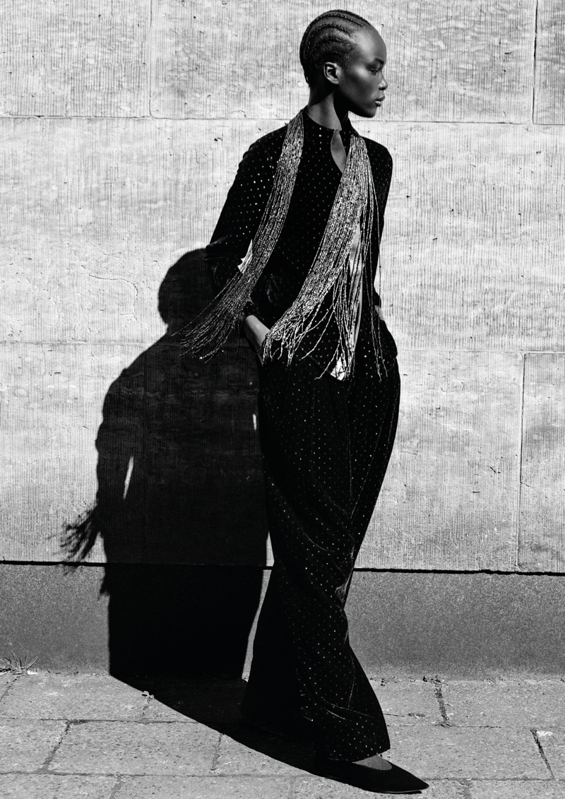Model standing outdoors in profile against a textured stone wall, casting a sharp shadow in bright sunlight.