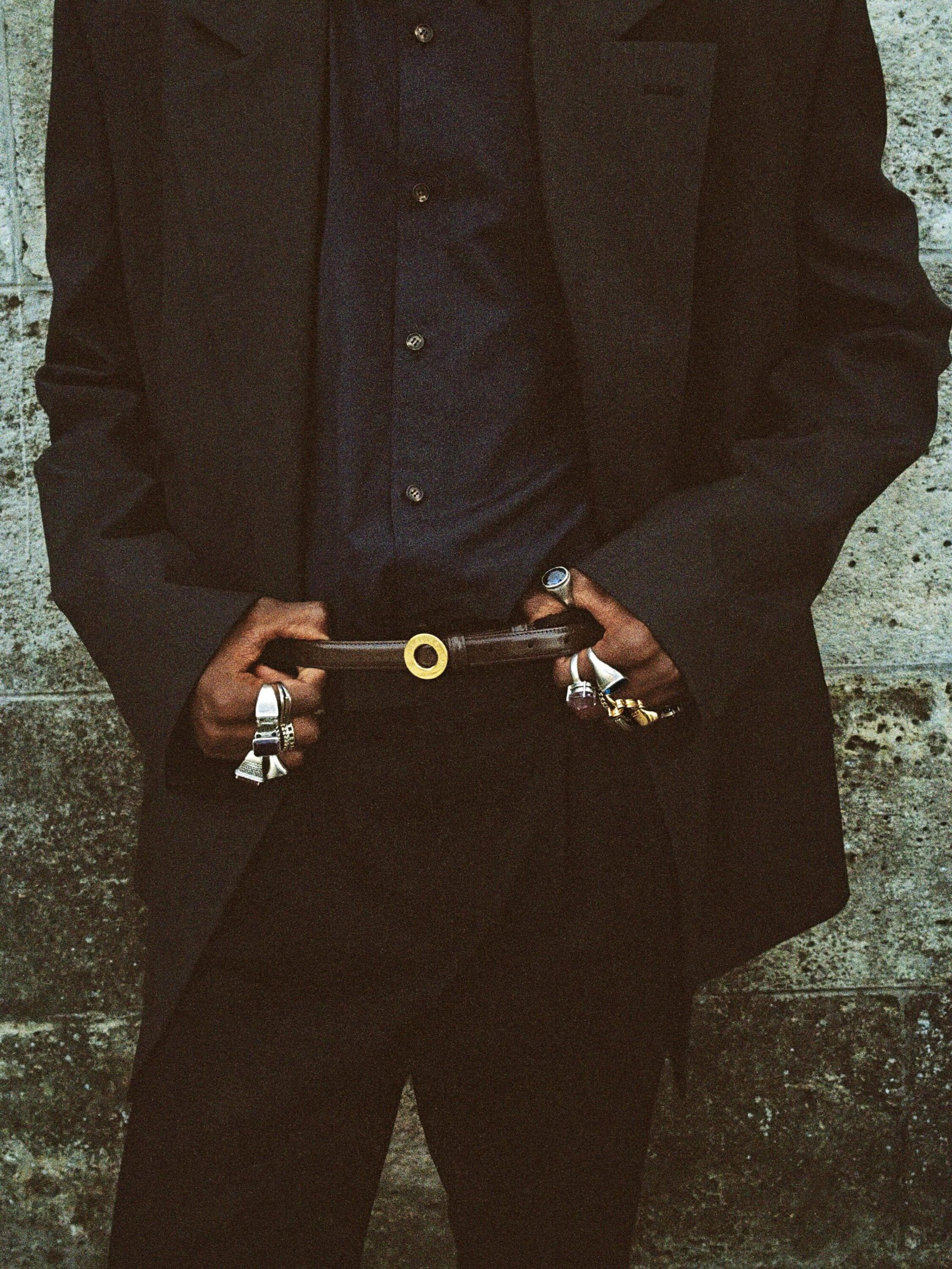 Close-up of a model wearing an oversized dark suit jacket, button-up shirt, and a thin belt with a gold ring buckle.