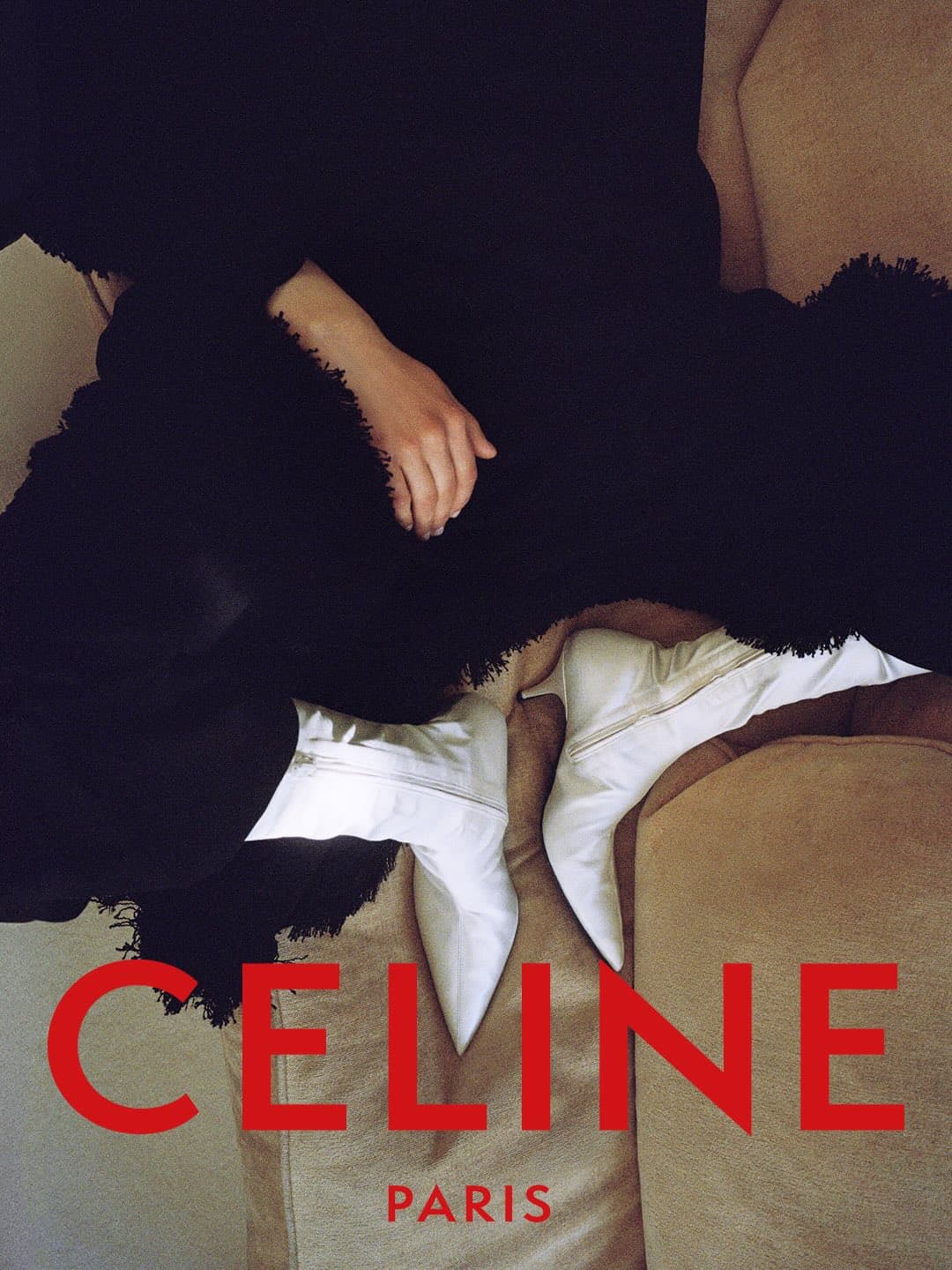 Person wearing pointed-toe white heeled ankle boots and a textured black garment on a sofa with red Celine Paris typography.