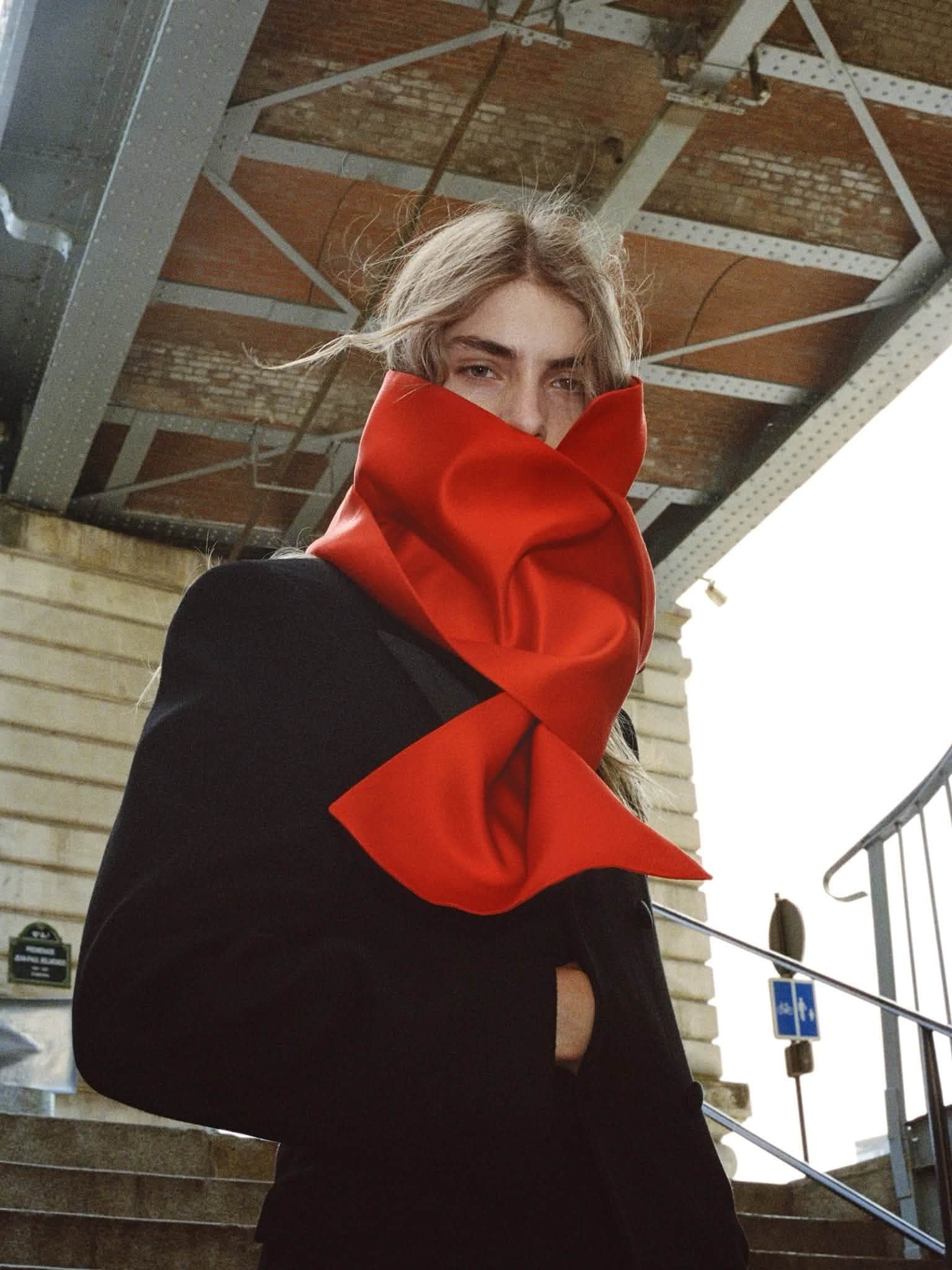 Model wearing a black coat and a structured oversized red scarf wrapped around the lower face under a bridge.