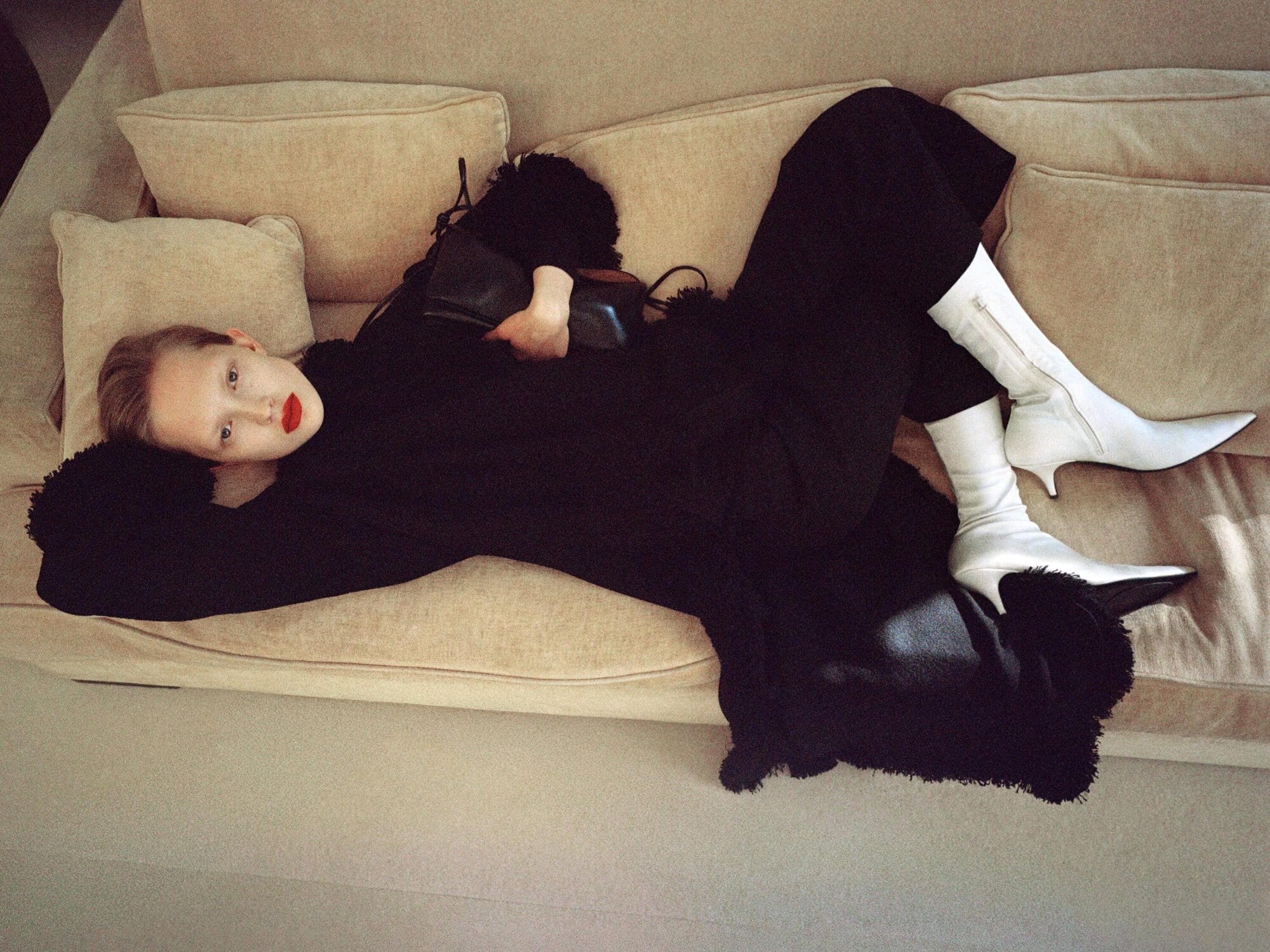 Model wearing a black outfit and white pointed-toe boots reclining horizontally on a beige sofa with bold red lips.