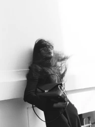 Model with windswept hair wearing dark glasses and holding a black leather handbag against a bright architectural ledge.