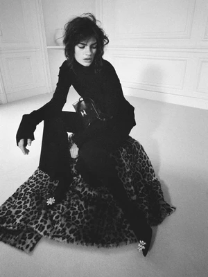 Model wearing an all-black outfit seated on a leopard print blanket in a classic paneled room, captured in high-contrast black and white.