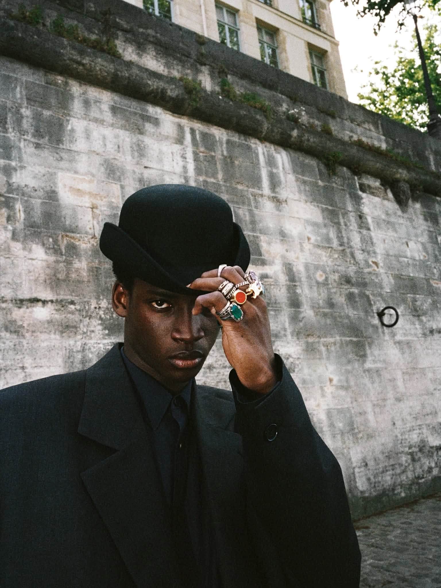 Black model wearing an all-black tailored suit and bowler hat with ornate gemstone rings against a stone building wall.