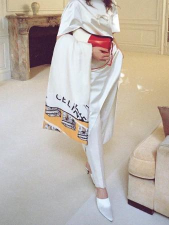 Model in an ivory silk outfit and pointed mules holding a red clutch and Celine printed scarf near an ornate fireplace.