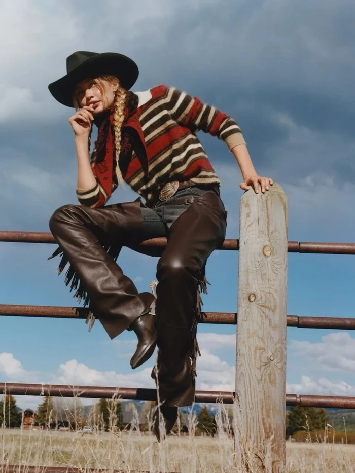 Model in cowboy hat and striped sweater perched on a wooden fence against an outdoor sky background