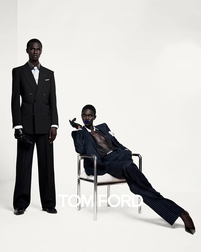 A man in a black suit standing next to a woman seated in a chrome chair wearing a pinstripe suit, against a white seamless background.