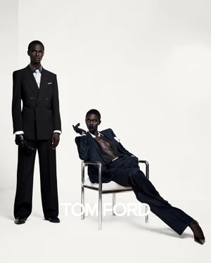 A man in a black suit standing next to a woman seated in a chrome chair wearing a pinstripe suit, against a white seamless background.