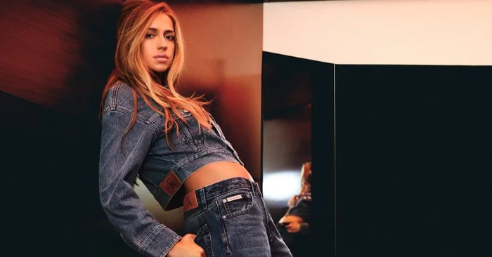 Woman in denim jacket and jeans leaning against a dark wall for Calvin Klein Feel the Fit