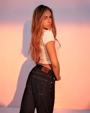 Woman with long blonde hair wearing a white top and dark jeans posing against a pink gradient studio background