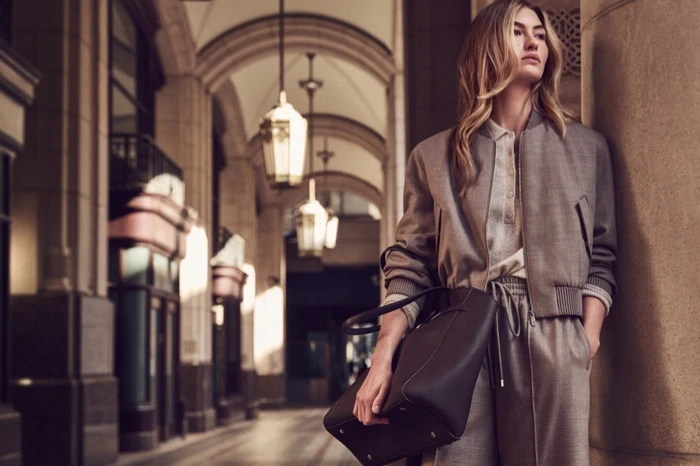Grace Elizabeth leaning against a column in a grey bomber jacket and matching trousers, holding a dark handbag.