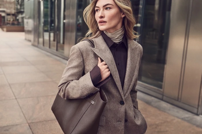 Grace Elizabeth wearing a tailored tweed blazer and carrying a dark leather tote bag outside a modern building.