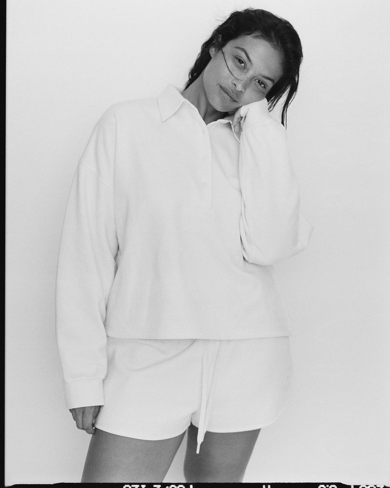 Woman in a white long-sleeved shirt and shorts poses against a white background in a studio shot. The image is in black and white.