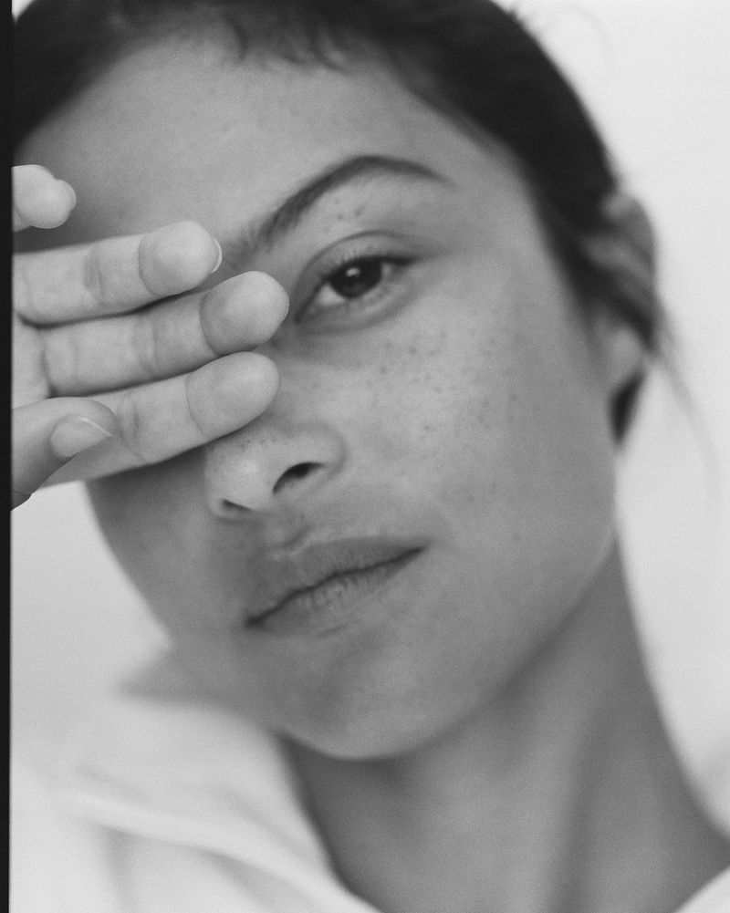Close-up black and white portrait of a young adult female with hand near face, set against a plain white background.
