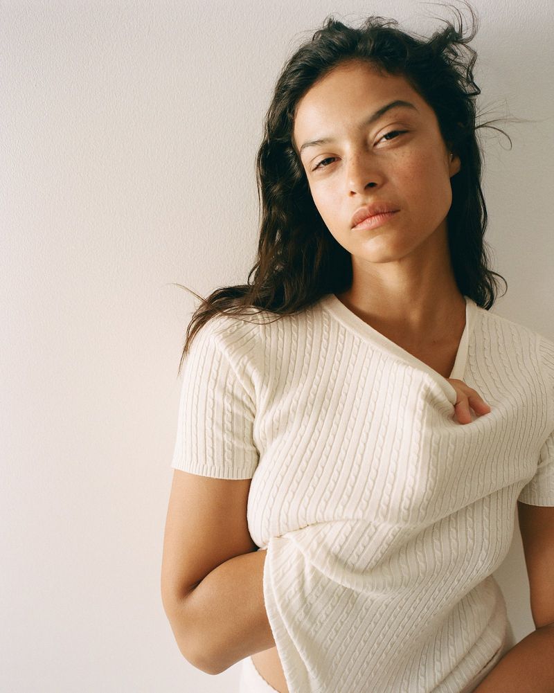 A woman with wavy brown hair poses indoors wearing a textured white V-neck knit shirt against an off-white backdrop in a portrait.