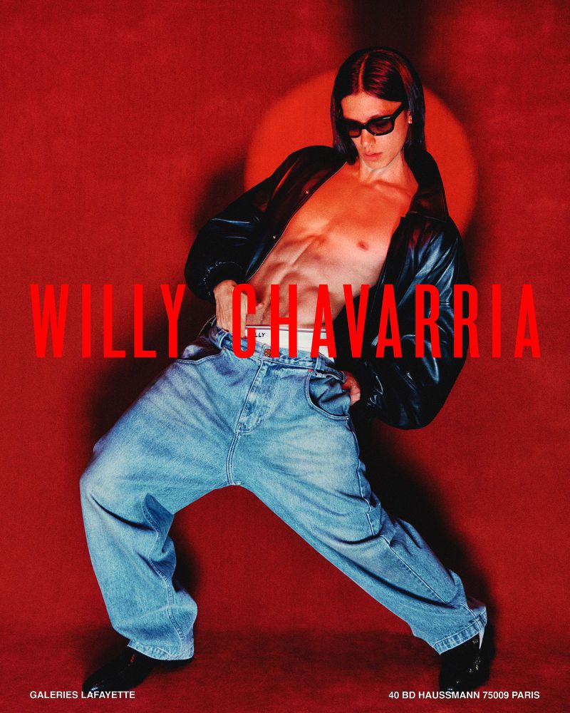 Shirtless male model in black leather jacket, baggy blue jeans, and dark sunglasses posing dynamically against a red backdrop.