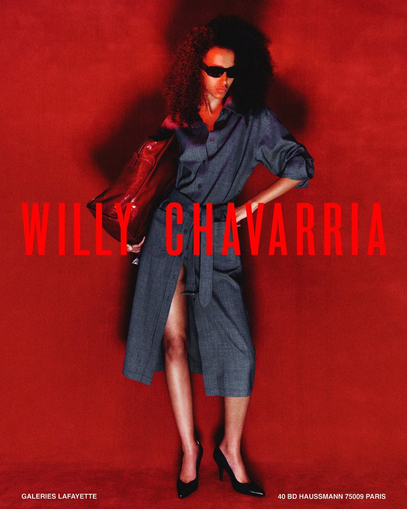Model wearing dark sunglasses and a grey belted shirt dress holding an oversized red bag against a deep red backdrop.