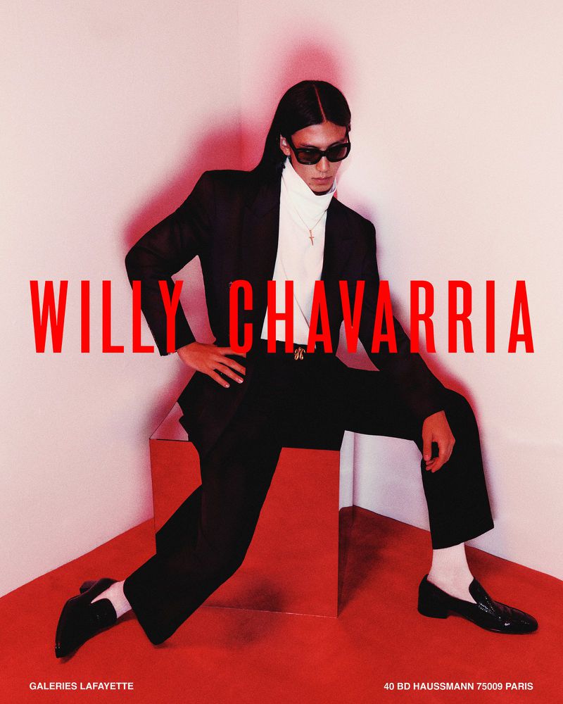 Model wearing a black suit, white turtleneck, and sunglasses seated on a reflective cube against a red-lit corner.