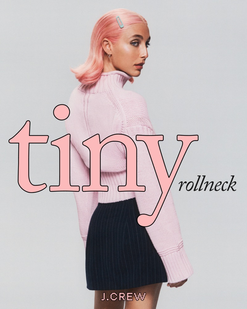 Emma Chamberlain wearing a light pink rollneck sweater and pinstripe miniskirt against a seamless grey backdrop.