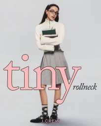 Emma Chamberlain wearing white zip-up sweater and grey pleated skirt holding books against light grey background for J. Crew