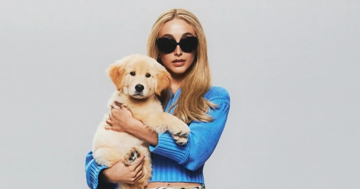 Emma Chamberlain wearing sunglasses and a blue sweater while holding a golden retriever puppy against a grey backdrop